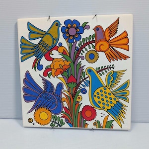 May include: A square, white ceramic tile featuring a colorful, folk art design. The artwork depicts three birds in shades of blue, yellow, and orange, perched amidst stylized flowers and foliage. The design is outlined in black, with a vibrant color palette.