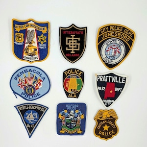 May include: A collection of nine police department patches. The patches vary in shape and design, featuring text such as "Metro Law Enforcement," "Interstate," and "City Police Dept." Colors include blue, gold, black, and white. Some patches display state seals and city names.