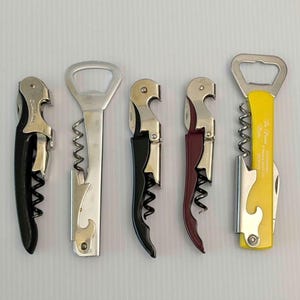 May include: Five waiter-style corkscrews arranged in a row. From left to right: a black handle, a plain silver metal finish, a black handle, a maroon handle, and a yellow handle with the text The Pioneer Woman printed in white script.