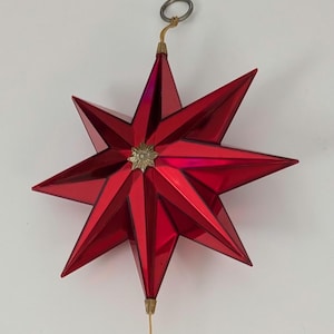 May include: A shiny red, multi-pointed star ornament with a metallic finish. The star features a small, gold-colored star-shaped emblem at its center. A thin gold wire extends from the bottom point, and a silver-colored metal ring is attached to the top.