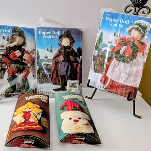 May include: Three Paper Doll Craft Kits in clear plastic packaging and two Patchwork Ornament Kits in rectangular boxes. The kits feature illustrations of dolls, an angel, and a Santa figure. The packaging displays text including Paper Doll Craft Kit.