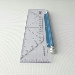 May include: A clear plastic parallel glider ruler featuring black printed markings, including a protractor scale and a 12-inch edge. The tool includes a blue metal cylindrical handle with a textured silver knob at the end, labeled Parallel Glider Israel.