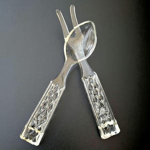 May include: Clear glass salad serving set with a fork and spoon. The handles have a diamond pattern with a floral design in the center. The fork has two tines. The spoon has a rounded bowl.