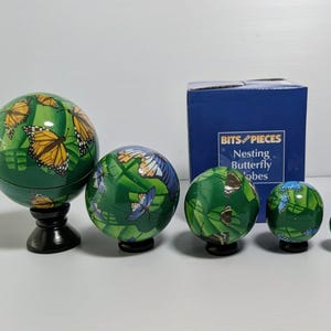 May include: A set of five green spherical nesting globes decorated with various butterfly illustrations, displayed alongside a blue product box labeled Bits and Pieces Nesting Butterfly Globes. The globes are arranged by size, with the largest resting on a black stand.