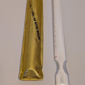 May include: A gold-colored, textured pouch with the text "MAGNER WITH RULER" next to a clear plastic tool with a ruler printed on it. The tool has a wide, flat end and a narrow, pointed end.