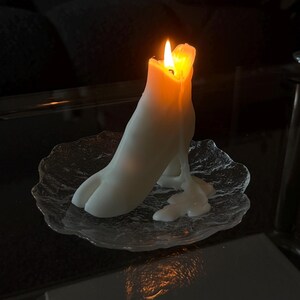 May include: A lit white candle shaped like a hoof, with melted wax dripping onto a clear, textured glass plate. The candle's flame glows brightly against the dark background, creating a warm ambiance. The candle is a unique decorative item.