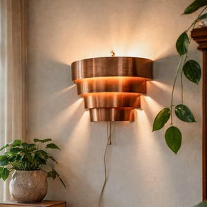 May include: A tiered copper wall sconce with a warm, metallic finish. The light fixture is mounted on a neutral-colored wall, casting a soft glow. A pull chain hangs down. A small potted plant and books sit on a wooden table.