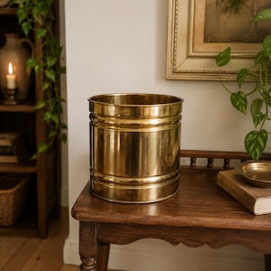 May include: A cylindrical brass planter with a polished finish. The planter has horizontal bands and vertical lines, and it sits on a wooden table. The planter is approximately 10 inches in diameter and 9 inches tall.