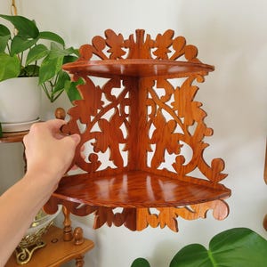 May include: A two-tier corner shelf made of polished brown wood with intricate cut-out scrollwork patterns. The unit features two triangular shelves connected by a decorative back panel that fits into a corner, showcasing a natural wood grain finish.