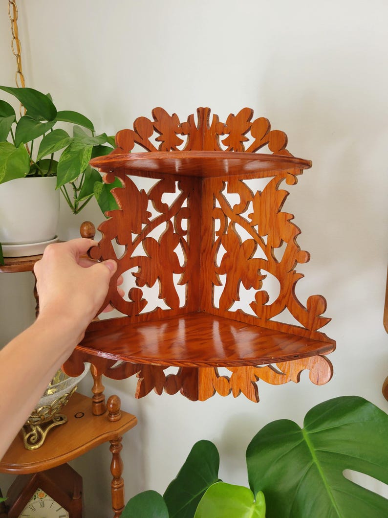 May include: A two-tier corner shelf made of polished brown wood with intricate cut-out scrollwork patterns. The unit features two triangular shelves connected by a decorative back panel that fits into a corner, showcasing a natural wood grain finish.