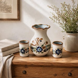 Peut inclure: Un vase en céramique à bord ondulé et deux tasses cylindriques assorties, tous dotés d'un émail crème moucheté avec des fleurs bleues, des feuilles vertes et un motif d'oiseau brun peint à la main sur le vase, posés sur une surface en bois.
