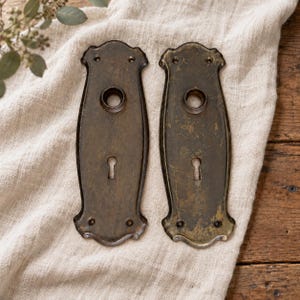 Peut inclure: Une paire de plaques de porte en laiton antique assorties avec une patine sombre et usée. Chaque plaque rectangulaire présente une ouverture circulaire centrale pour une poignée, une serrure en bas et quatre petits trous de vis.