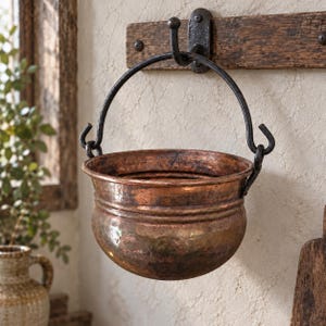 May include: A round, hammered copper pot with a dark metal handle, suspended from a black iron hook attached to a horizontal wooden plank. The copper vessel features a wide rim and a rounded body with a polished, metallic finish.