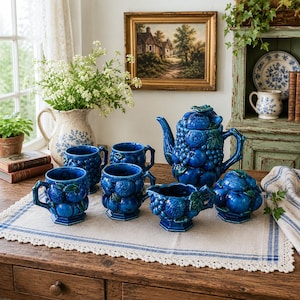 May include: A ceramic tea set featuring a deep blue glaze with a raised, three-dimensional fruit and leaf motif. The set includes a teapot with a lid, a creamer, a sugar bowl with a lid, and four mugs, all resting on a rectangular linen placemat.