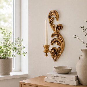 May include: A gold-coloured wall-mounted candle sconce featuring an ornate, swirling acanthus leaf design. The sconce holds a single, slender, off-white taper candle in a small, rounded cup extending from the lower section of the decorative frame.