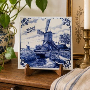 May include: A square ceramic tile with a blue and white windmill scene, with the word "Holland" inscribed. The tile is displayed on a wooden stand. The image evokes a sense of Dutch heritage and craftsmanship.