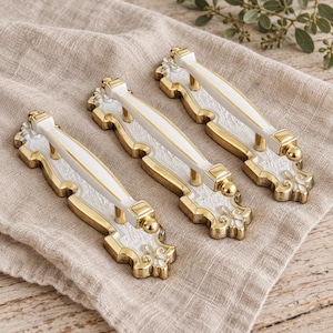 Peut inclure: Trois poignées de tiroir composées d'une barre centrale blanche avec des accents dorés, montées sur une plaque décorative dorée et blanche avec des détails en relief complexes et un motif de fleur de lys à chaque extrémité.