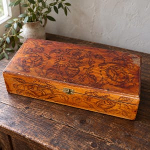 May include: A rectangular wooden box with a warm brown finish, featuring dark, intricate pyrography designs of floral patterns and a central vase motif on the lid. The box includes a small, centred brass-coloured metal latch on the front edge.