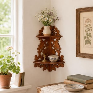 May include: A two-tiered wooden corner shelf featuring intricate carved scrollwork patterns. The top shelf holds a small cream-colored ceramic vase filled with white daisies and wildflowers, while the bottom shelf displays a brass candlestick and a small bowl.