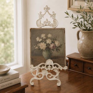 May include: A square painting of a floral bouquet in a vase displayed on an ornate, distressed white metal easel. The artwork features white and soft pink flowers in a light green vase against a muted, dark background, held by a stand.