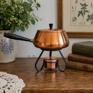 May include: A copper fondue pot with a black handle and a matching lid featuring a black knob. The pot rests on a black metal stand that includes a small copper burner underneath. The design is metallic and functional with a polished finish.