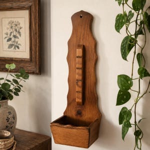 May include: A wooden wall-mounted planter, featuring a rectangular box base and an elongated backboard with a decorative edge. The backboard has a series of small, rectangular holders. The planter is stained a warm, medium brown colour, and is designed for wall mounting.