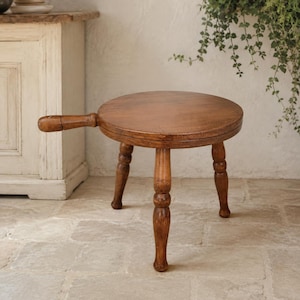 May include: A round wooden stool with a smooth, dark brown finish and three turned legs. The stool features a single horizontal handle extending from one side of the circular seat, resembling a paddle or a small serving board with legs.