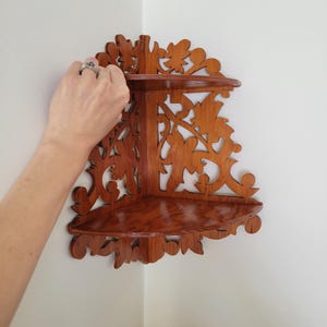 May include: A two-tier wooden corner shelf featuring a rich brown finish and intricate, laser-cut floral scrollwork patterns along the back panels. The unit consists of two triangular shelves designed to fit flush against a wall corner.