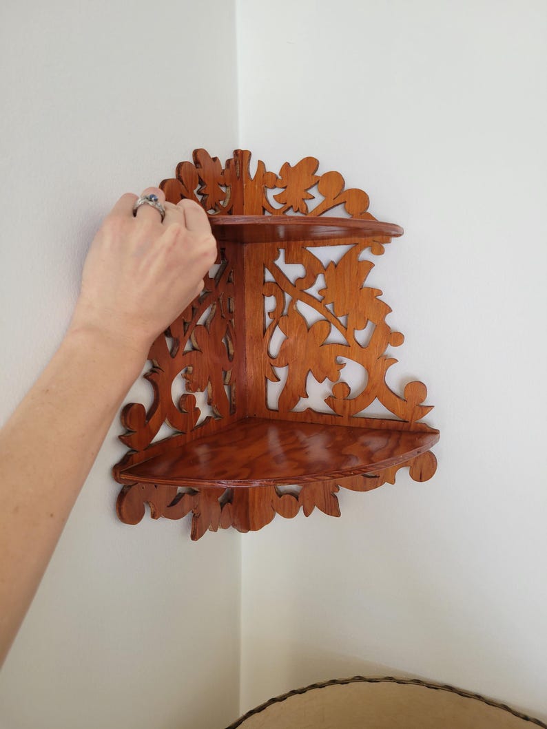 May include: A two-tier wooden corner shelf featuring a rich brown finish and intricate, laser-cut floral scrollwork patterns along the back panels. The unit consists of two triangular shelves designed to fit flush against a wall corner.