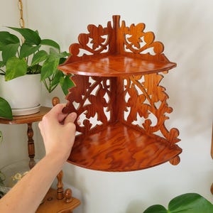 May include: A two-tier corner shelf made of polished brown wood with a visible grain pattern. The shelf features intricate, openwork scroll and leaf carvings along the side panels and the top back edge, designed to fit into a 90-degree corner.