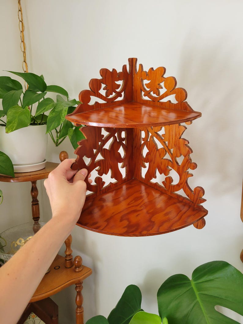 May include: A two-tier corner shelf made of polished brown wood with a visible grain pattern. The shelf features intricate, openwork scroll and leaf carvings along the side panels and the top back edge, designed to fit into a 90-degree corner.