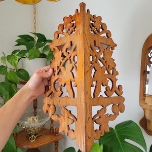 May include: A light brown wooden corner shelf featuring intricate, symmetrical cut-out scrollwork and leaf-like patterns. The shelf has a vertical central spine and a flat top surface, designed to fit into a corner with a natural wood grain finish.
