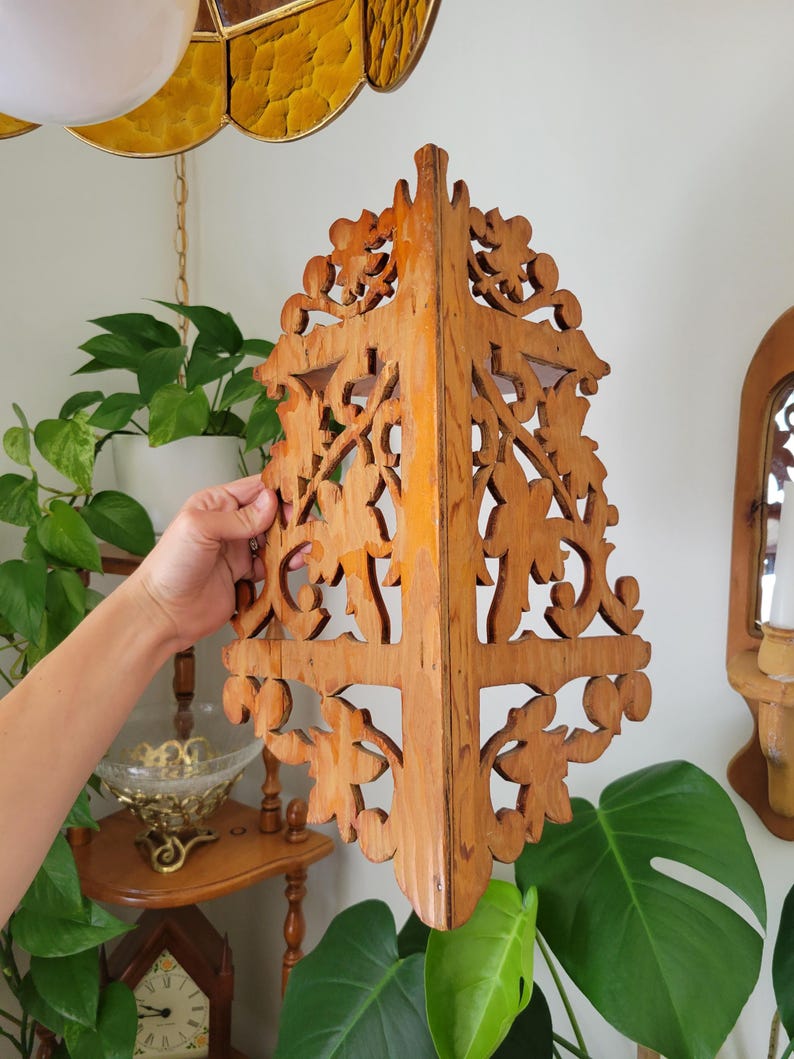 May include: A light brown wooden corner shelf featuring intricate, symmetrical cut-out scrollwork and leaf-like patterns. The shelf has a vertical central spine and a flat top surface, designed to fit into a corner with a natural wood grain finish.