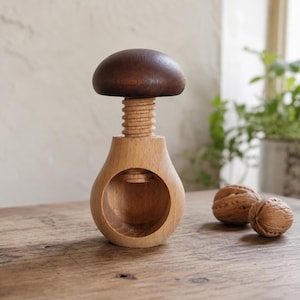 Peut inclure: Un casse-noix en bois en forme de champignon avec un chapeau brun foncé et une base beige clair. La partie supérieure comporte un mécanisme à vis fileté qui s'ajuste verticalement à l'intérieur du centre évidé du corps en bois clair.