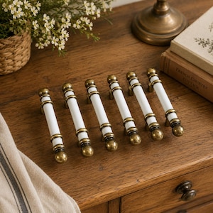Peut inclure: Un ensemble de six poignées de tiroir, chacune composée d'une section centrale cylindrique en céramique blanche, agrémentée de garnitures en métal couleur laiton antique aux extrémités et au centre, avec des embouts sphériques.