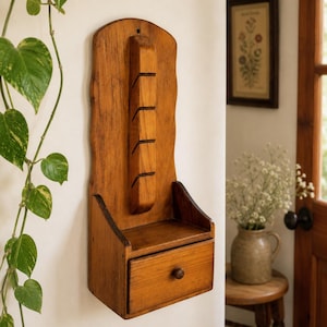 May include: A wooden wall-mounted organiser with a drawer and several slots. The organiser is made of light brown wood and has a rustic, handcrafted appearance. It is designed to hold small items and is mounted on a white wall.