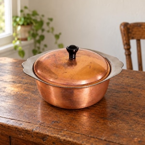 May include: A round copper cooking pot with a matching lid and a black knob handle. The pot features wide, scalloped silver-toned metal side handles. The copper surface shows natural variations in colour and texture throughout the exterior.