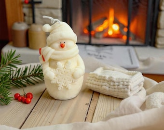 May include: A snowman-shaped candle with a hat, scarf, and snowflake detail. The candle is white with a red nose and scarf accents. A small, white, rectangular, knitted item is next to the candle. The background includes a fireplace and wooden surface.