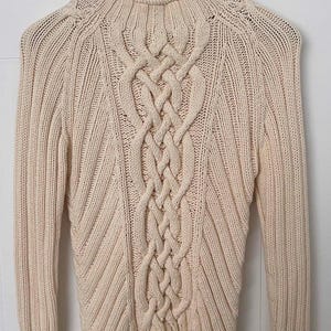 May include: A cream-coloured long-sleeve jumper featuring a prominent central vertical cable knit pattern flanked by diagonal ribbed textures. The garment has a high mock neck collar and is displayed on a wooden hanger against a white background.
