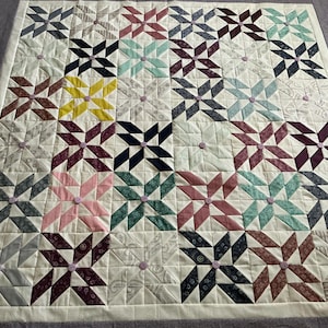May include: A handmade quilt featuring a repeating starburst pattern in various colors, including navy, teal, pink, and yellow, set against a cream background. The quilt is bordered with a gray fabric and has small, light purple buttons at the center of each star.
