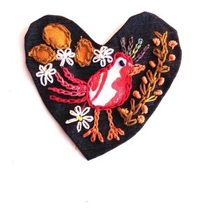 May include: Embroidered heart-shaped patch featuring a red and white bird with floral and leaf accents. The bird has a white chest and red tail feathers. The patch is on a dark grey background.