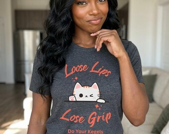 May include: A dark heather gray short-sleeve t-shirt featuring a graphic of a white cat with orange stripes peeking over a line. The text reads Loose Lips Lose Grip and Do Your Kegels in orange lettering with small red star accents.