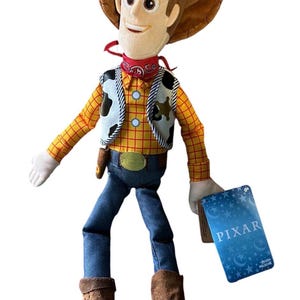 Op de afbeelding: Een pluche pop van een cowboyfiguur met een bruine hoed, een geel en rood geruit overhemd, een vest met koeienprint, een blauwe broek en bruine laarzen met sporen. Een rode bandana zit om de nek en een blauw label met PIXAR hangt aan de hand.