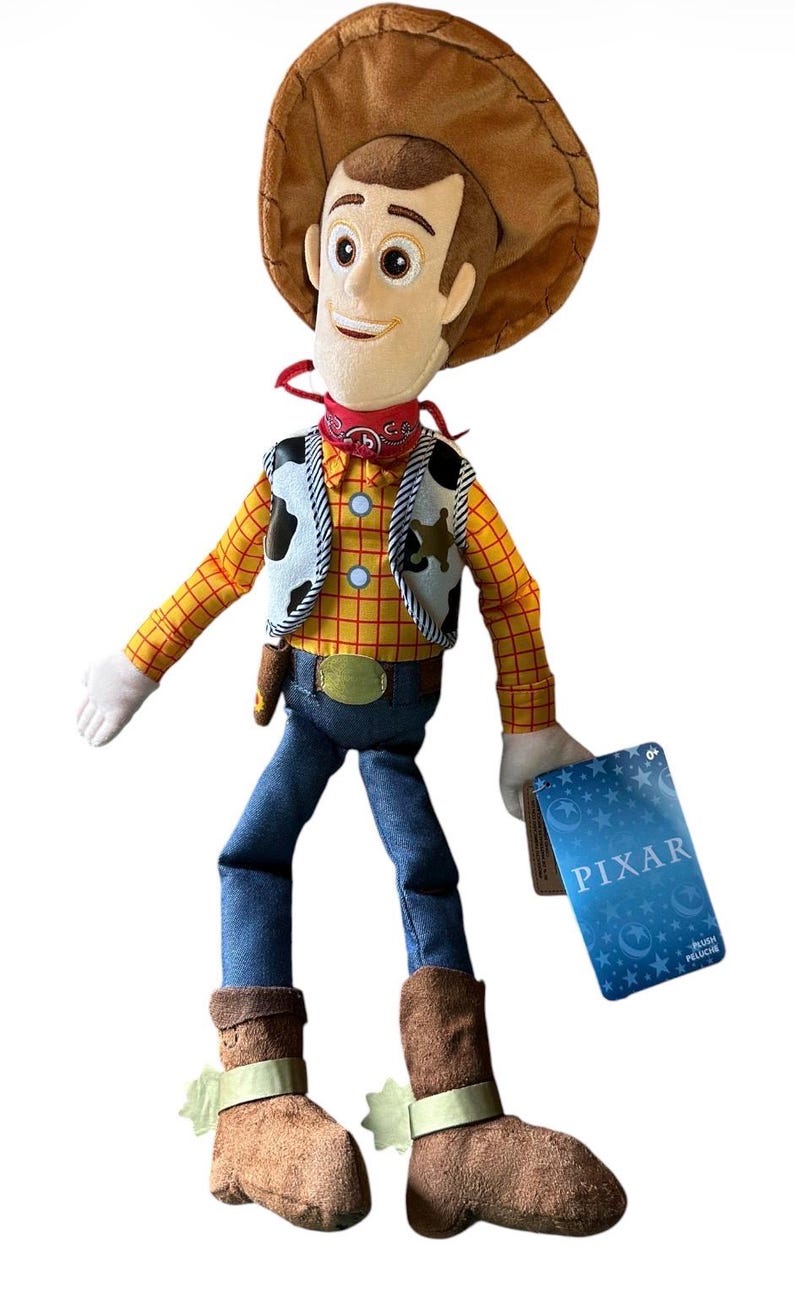 Op de afbeelding: Een pluche pop van een cowboyfiguur met een bruine hoed, een geel en rood geruit overhemd, een vest met koeienprint, een blauwe broek en bruine laarzen met sporen. Een rode bandana zit om de nek en een blauw label met PIXAR hangt aan de hand.