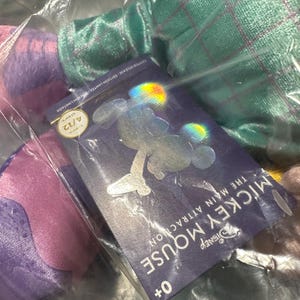 May include: A Disney Mickey Mouse The Main Attraction series item, visible through plastic packaging. The tag shows a Mickey Mouse silhouette and the text "MICKEY MOUSE THE MAIN ATTRACTION" and "4/12". The item is in a clear plastic bag.