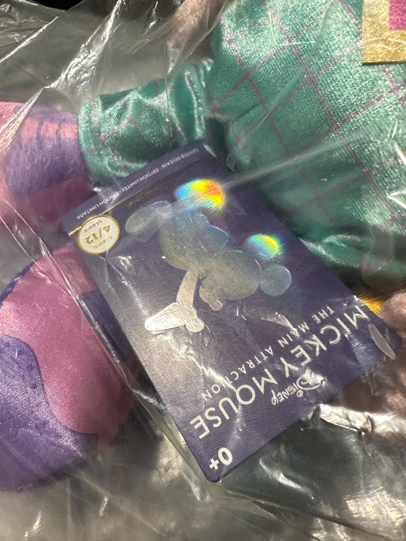 May include: A Disney Mickey Mouse The Main Attraction series item, visible through plastic packaging. The tag shows a Mickey Mouse silhouette and the text "MICKEY MOUSE THE MAIN ATTRACTION" and "4/12". The item is in a clear plastic bag.