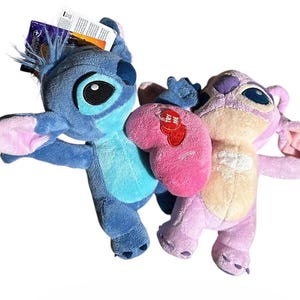 May include: Two plush toys featuring blue and pink characters with large ears, dark eyes, and light-colored chest patches. The blue character has a tag attached to its head, and the pink character holds a bright pink heart with a red Try Me tag.
