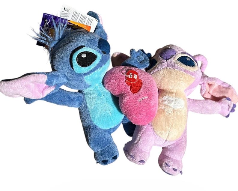 May include: Two plush toys featuring blue and pink characters with large ears, dark eyes, and light-colored chest patches. The blue character has a tag attached to its head, and the pink character holds a bright pink heart with a red Try Me tag.