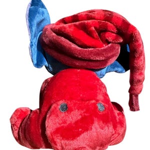 May include: A plush toy featuring a bright red body with a soft texture and two small dark circular eyes. The toy is topped with a long, drooping red hat that curves downward, accented by a blue fabric element visible behind the red hat section.