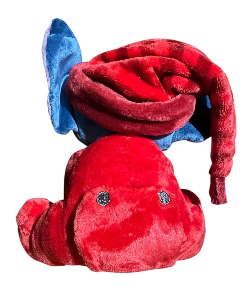 May include: A plush toy featuring a bright red body with a soft texture and two small dark circular eyes. The toy is topped with a long, drooping red hat that curves downward, accented by a blue fabric element visible behind the red hat section.